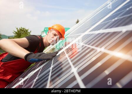 Ingenieur Inspektion von Solaranlagen in der Nähe des Hauses. Nahaufnahme. Der Mann trägt an einem warmen Tag eine orangefarbene Uniform, einen harten Hut, ein schwarzes T-Shirt und Handschuhe. Blaue Tafeln reflektieren die Sonnenstrahlen Stockfoto