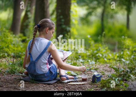 Mädchen Maler malt Bäume im Park. Mädchen in Denim Overalls sitzen mit ihrem Rücken Stockfoto