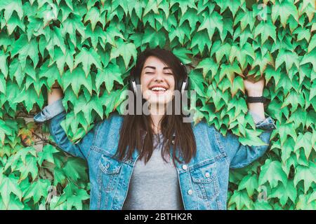 Junge Hipster-Mädchen hört Musik, die sich an eine Efeu-Wand lehnt - hübsche Frau entspannt sich mit Kopfhörern in einem Stadtpark Stockfoto