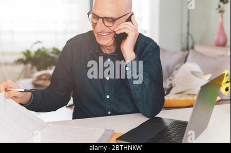 Mann, der am Telefon spricht und lacht, während er Arbeitsdokumente überprüft - Reife Mann, der von zu Hause aus am Computer arbeitet - Fernarbeitskonzept Stockfoto