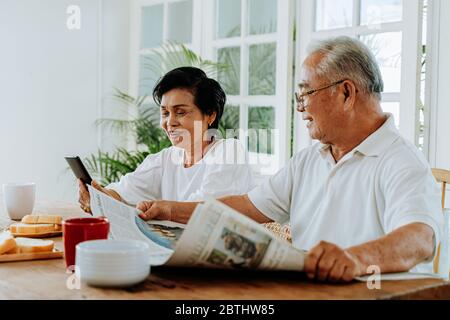 Älteres asiatisches Paar, das zusammen im Esszimmer frühstückte. 70er Jahre Rentner älterer Mann liest Zeitung, während Frau mit Handy Stockfoto