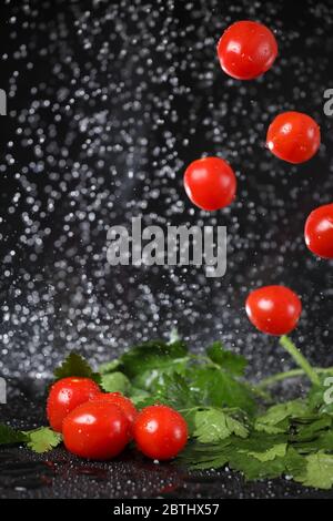 Vertikale Ansicht der fallenden roten Kirschtomaten unter Wassertropfen in einem schwarzen Hintergrund. Vegetarisches Essen. Gesunde Lebensweise. Sommerdiät Stockfoto