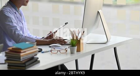 Beschnitten Bild von jungen Arzt mit einem Klemmbrett in der Hand während der Arbeit und sitzt vor Computer-Monitor, dass auf weißen Schreibtisch setzen, dass Stockfoto