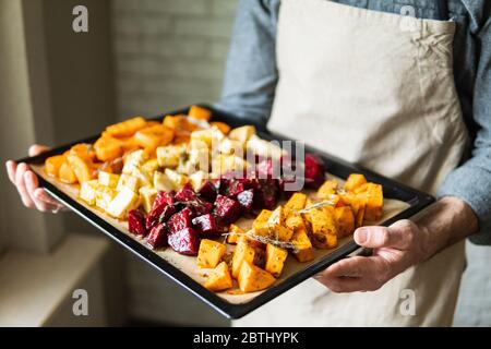 Männliche Hände halten Backblech mit Gemüse mischen. Vegetarier kochen Süßkartoffeln und andere Gemüse zu Hause. Stockfoto