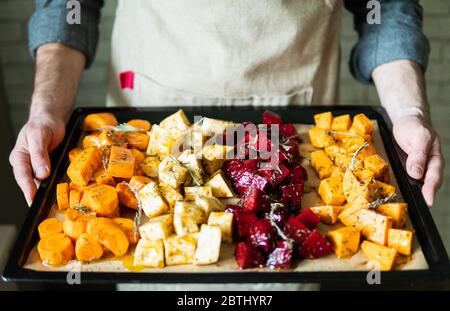 Männliche Hände halten Backblech mit Gemüse mischen. Vegetarier kochen Süßkartoffeln und andere Gemüse zu Hause. Stockfoto
