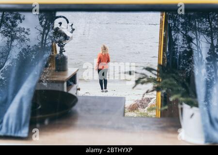 Junge blonde Frau neben dem Wohnmobil mit Blick auf das gefrorene Wintermeer. Blick durch die offene Tür. Stockfoto