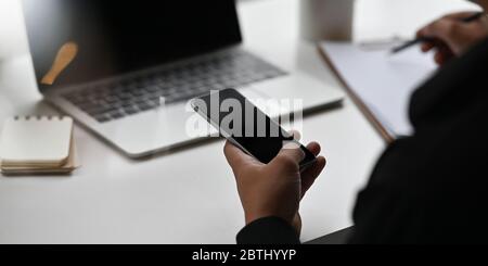 Abgeschnitten von Geschäftsleuten die Hände halten ein Smartphone während Schreiben auf Zwischenablage und sitzen vor Computer-Laptop, dass auf weißen Schreibtisch setzen Stockfoto