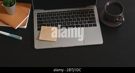 Foto von Computer-Laptop auf schwarzen Schreibtisch setzen und umgeben von Keramik Kaffeetasse, Stapel von Notebook und Stift. Geordnetes Arbeitsplatzkonzept. Stockfoto