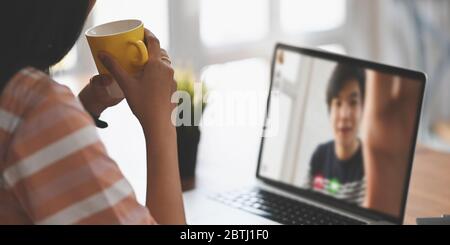 Bild abgeschnitten von schönen Frau, die einen heißen Kaffee trinkt, während sie vor ihrem Computer-Laptop sitzt, das Video, das mit ihrem Freund über auf dem Bildschirm anruft Stockfoto