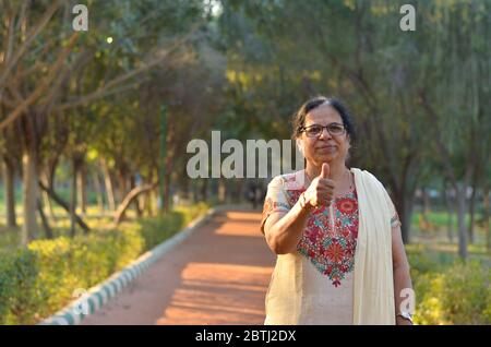 Porträtaufnahme einer glücklich aussehenden älteren nordindischen Frau, die traditionellen Chikan kari indischen Salwar Kameez trägt, der einen Daumen nach oben in einem Garten zeigt Stockfoto