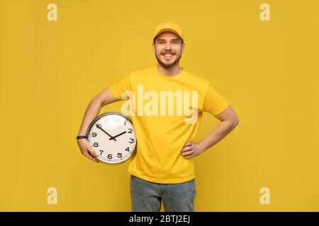 Lieferung Mann in gelb Arbeitskleidung halten Uhr isoliert auf gelben Hintergrund Studio Porträt. Stockfoto