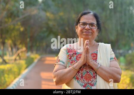 Smart Senior nordindische Frau stehend, posiert für die Kamera mit Händen in namaste gefaltet als Zeichen des Respekts und willkommen in einem Park tragen ab wh Stockfoto