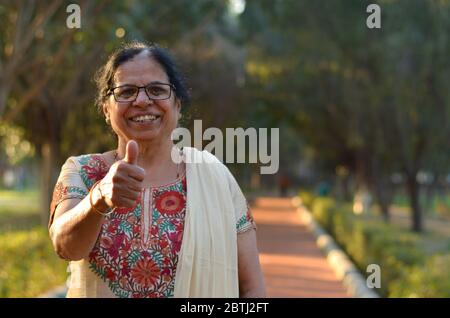 Porträtaufnahme einer glücklich aussehenden älteren nordindischen Frau, die traditionellen Chikan kari indischen Salwar Kameez trägt, der einen Daumen nach oben in einem Garten zeigt Stockfoto