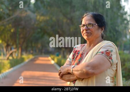 Intelligente und selbstbewusste ältere nordindische Frau stehend, denken und wegschauen mit gekreuzten / gefalteten Händen in einem Park trägt eine off weißen Salwar ka Stockfoto