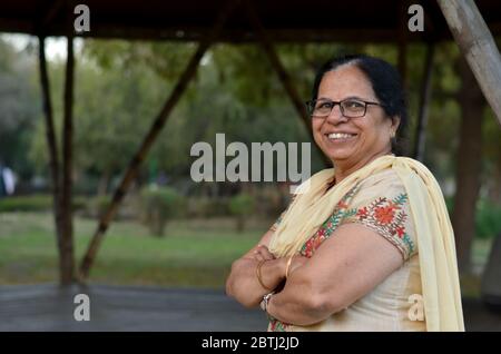 Intelligente und selbstbewusste ältere nordindische Frau stehend, posiert für die Kamera mit gekreuzten / gefalteten Händen in einem Park trägt einen off weißen Salwar Kameez Stockfoto