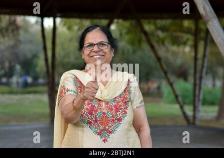 Porträtaufnahme einer glücklich aussehenden älteren nordindischen Frau, die traditionellen Chikan kari indischen Salwar Kameez trägt und einen Daumen nach oben in einem Garten zeigt Stockfoto