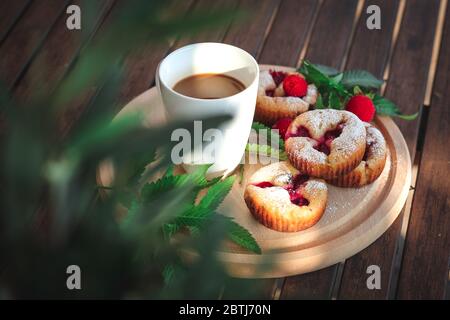 Cupcakes mit Beeren und einer Tasse Kaffee auf einem dunklen Gartentisch, grüne Blütenblätter in Bokeh Stockfoto