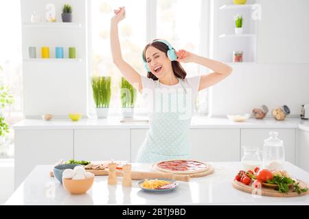 Porträt von fröhlichen funky Mädchen Hausfrau kochen Pizza Stellen Sie sich vor, sie Pop Start haben Ruhe entspannen hören Headset Musik Melodie singen tragen gepunktete Schürze im Haus Stockfoto