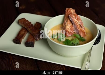 Hausgemachte dicke Erbsensuppe mit geräucherten Schweinerippen in einer tiefen Keramikschale auf einem dunklen Holztisch. Ein Zweig von Grün. Roggenbrot Croutons auf einer Platte. Sou Stockfoto