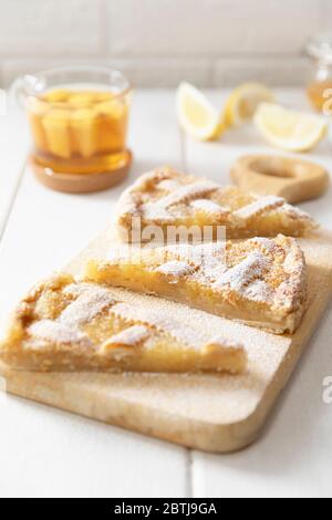 Drei Stücke von hausgemachten Mürbbrot Zitronenkuchen mit Puderzucker auf einem Schneidebrett bestreut. Tee in einem Becher und Zitronenscheiben im Hintergrund. Stockfoto