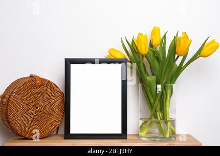 Leerer Rahmen auf einem kleinen Holztisch mit gelben Tulpen und Strohbeutel Stockfoto
