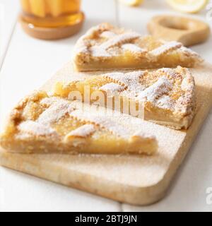 Drei Stücke von hausgemachten Mürbbrot Zitronenkuchen mit Puderzucker auf einem Schneidebrett bestreut. Tee in einem Becher und Zitronenscheiben im Hintergrund. Stockfoto