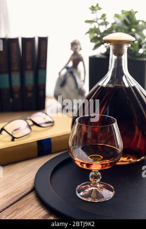 Cognac in einem Schnitter und Flasche auf einem schwarzen Tablett auf einem Holztisch. Bücher, Gläser, eine Blume und eine Porzellanfigur im hellen Licht des Windo Stockfoto