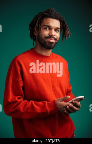 Porträt eines jungen afrikanischen Mannes mit Pullover isoliert auf grünem Hintergrund stehend, mit Handy Stockfoto
