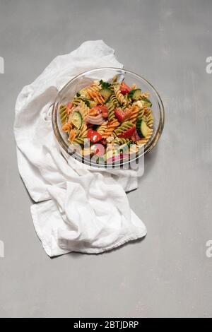 Glasschüssel voll mit Nudelsalat mit Fusilli, Feta-Käse, Gemüse und Olivenöl Dressing. Weißes Tuch auf der Seite, und grau verzweifelt Hintergrund. ref Stockfoto