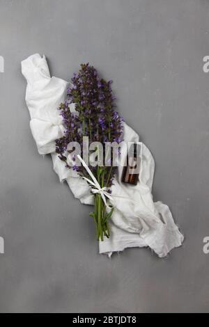 Dunkle bernsteinfarbene Glasflasche voller ätherischer Öl und Salbeiblüten, Salvia sclarea, für Aromatherapie flach auf weißem Tuch und grauem Hintergrund legen Stockfoto