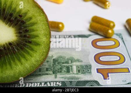 Erschwingliches Konzept für frische gesunde Lebensmittel: Nahaufnahme von frischen Kiwi, gelben Nahrungsergänzungskapseln und 100-Dollar-Banknote (Schwerpunkt Kiwi) Stockfoto