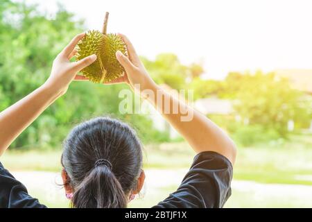 Junge Frau trägt eine Schutzmaske, um eine Infektion des Corona-Virus (Covid-19) in einem schwarzen Hemd zu verhindern, das einen reifen Durian über ihrem Kopf hält, New Normal li Stockfoto