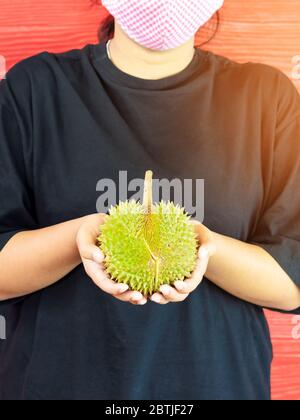 Junge Frau trägt eine Schutzmaske, um eine Infektion des Corona-Virus (Covid-19) in einem schwarzen Hemd mit einem reifen Durian zu verhindern, bereit, mit dem roten w zu essen Stockfoto