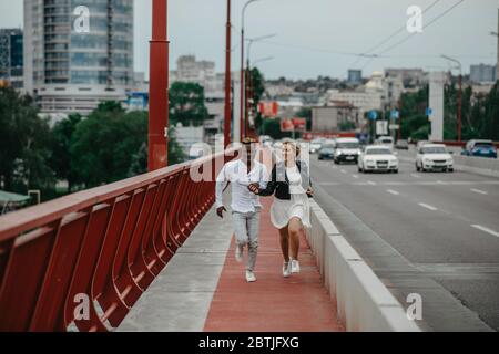 Junge interracial Paar hält die Hände, läuft auf Brücke und lacht fröhlich auf dem Hintergrund der Stadt und der Straße. Das Konzept der Liebe relationshi Stockfoto