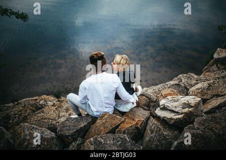 Interracial Paar sitzt auf Felsen und Umarmungen vor dem Hintergrund des Flusses. Konzept der Liebesbeziehungen und Einheit zwischen verschiedenen menschlichen Rassen. Stockfoto