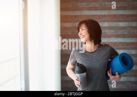 Erwachsene fit schlanke Frau hat Training zu Hause. Fröhliche positive ältere weibliche Person lächelnd und Blick auf das Fenster. Frau nach dem Training trinken Protein Stockfoto