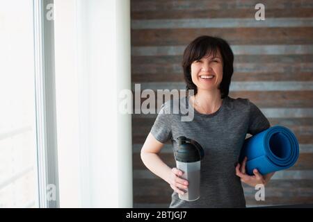 Erwachsene fit schlanke Frau hat Training zu Hause. Positive fröhliche ältere Frau schaut auf Kamera und Lächeln. Hält Protein Shake und Yoga Matte in den Händen Stockfoto