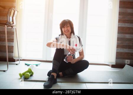 Erwachsene fit schlanke Frau hat Training zu Hause. Senior fröhlich attraktives Modell lächelnd vor der Kamera. Auf Yogamatte sitzen und Wasserflasche halten. Pause während Stockfoto