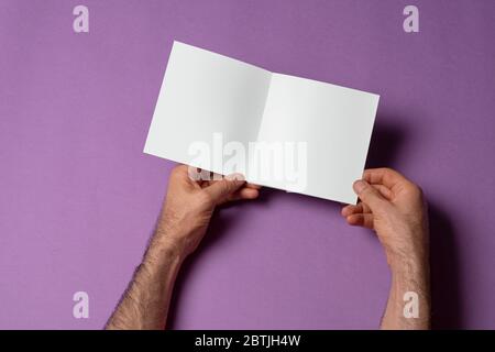 Männliche Hände halten eine geöffnete quadratische Größe dreifach-Katalog auf lila Hintergrund, Mock-up-Serie Vorlage bereit für Ihre Design-Auswahl-Pfad enthalten Stockfoto