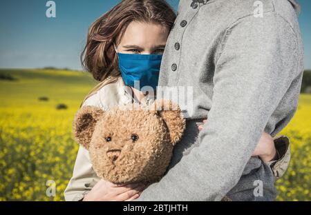 Kaukasische Mädchen tragen Blue Safe Breathing Maske Umarmung ihr Vater und Teddybär während Pandämie. Landschaftlich schöne sonnige Landschaft im Hintergrund. Alles Gute, Familie Tim Stockfoto