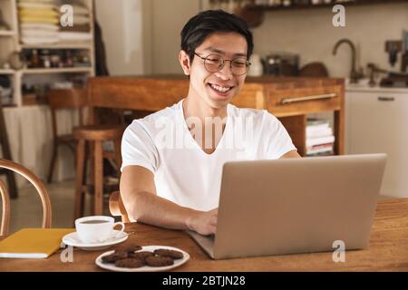 Bild von asiatischen glücklichen Mann lächelnd und mit Laptop, während am Tisch in der gemütlichen Küche sitzen Stockfoto