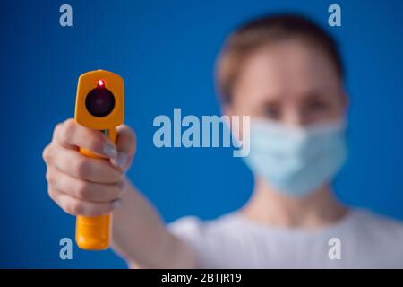 Frau in medizinischer Gesichtsmaske mit gelbem Pyrometer zur Temperaturmessung in Richtung Kamera - Nahaufnahme, selektiver Fokus. Gesundheitswesen, Messung Stockfoto