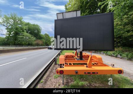 Ein leeres digitales Autobahnschulterschild, für Kopierraum, mit der Straße auf einer Seite und einem Auto, das in der Ferne wegfährt, umgeben von Laub. Stockfoto