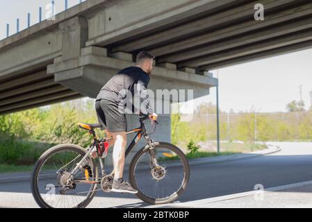 Radfahrer, die auf offener Straße ein Fahrrad fahren, um den Sonnenuntergang-Sportler, bärtigen Radler, Fahrten auf der Autobahn auf einem Sportfahrrad. Unter der Brücke. Im Freien. S Stockfoto