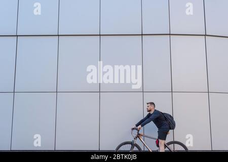 Bärtiger Radfahrer auf einem Fahrrad auf grauem Gebäudehintergrund. Im Freien. Frühlingssport. Der Typ ist fünfundzwanzig Jahre alt. Radfahren Fahrradtour im Frühling. Extr Stockfoto