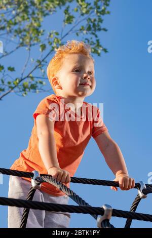 Kleiner Junge, der auf einem Spielplatz auf Seilen steht und selbstbewusst in die Ferne schaut. Ein neuer Wachstumszweig... Stockfoto