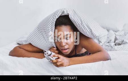 Überschlaf-Konzept. Schockiert afroamerikanischen Mädchen wacht auf, klettert aus unter Abdeckungen Blick auf Wecker Stockfoto
