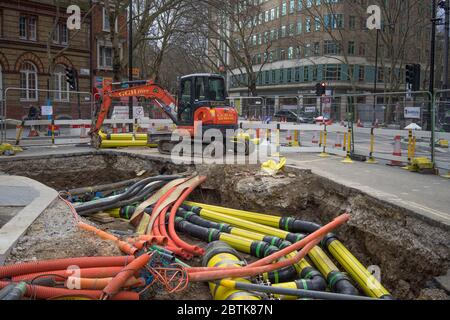 Straßenarbeiten mit einem Bagger an der Seite eines großen Lochs mit vielen Wasser-, Gas- und Stromleitungen. Ecke High Holborn und Shaftesbury Ave Stockfoto