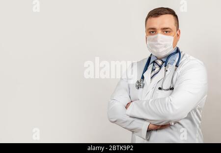 Ein Arzt in einer medizinischen Maske mit gekreuzten Armen, auf der Brust posiert während der COVID-19 in einem weißen Mantel vor der Kamera in der Nähe eines weißen Gebäudes. Stockfoto