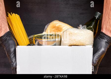 Nahaufnahme mans Hände in schwarzen Gummihandschuhen hält eine weiße Box mit Pasta, Getreide und Konserven auf schwarzem Hintergrund. Sicherheit Lebensmittel Lieferservice Stockfoto